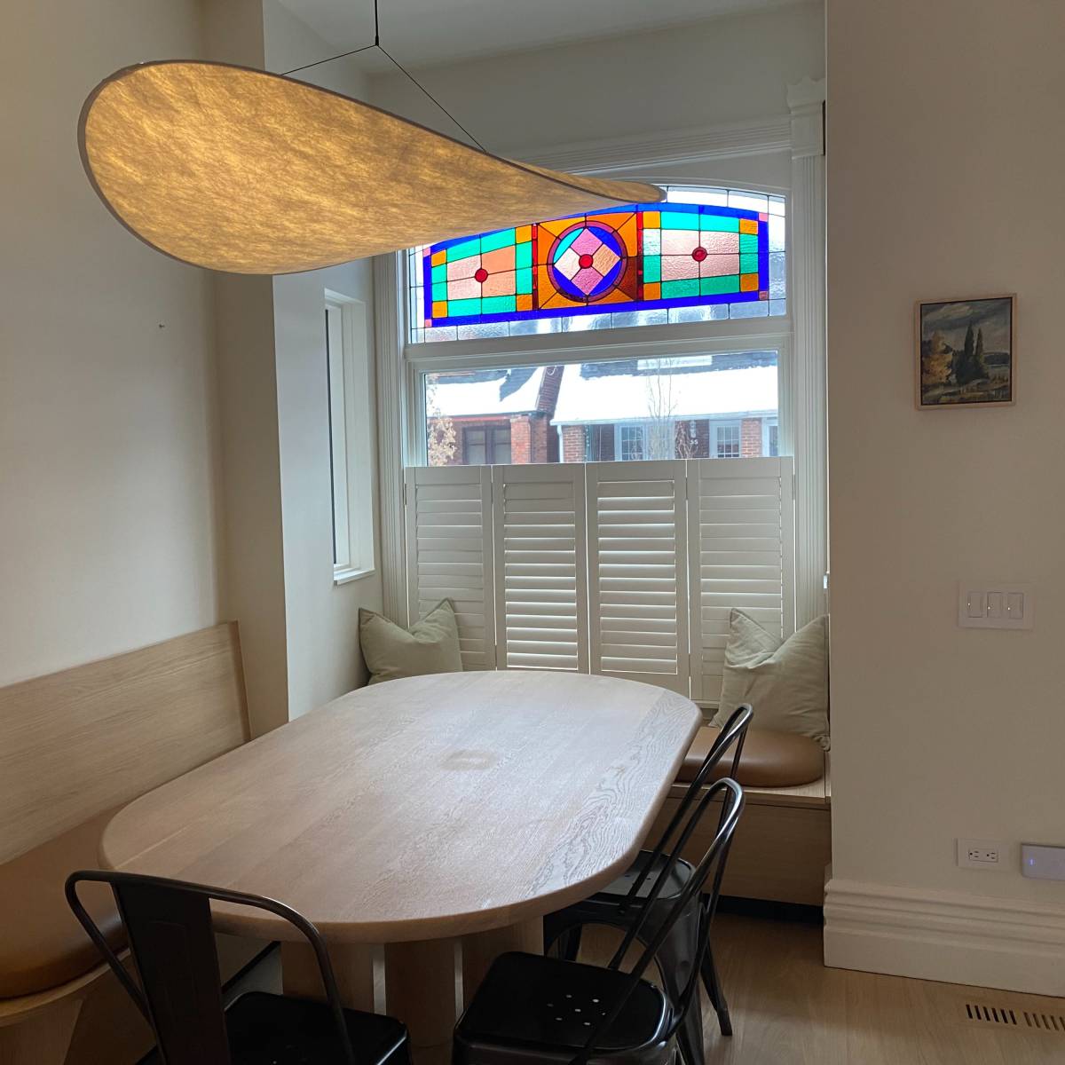 A modern dining area features a large, oval wooden table surrounded by black chairs, with a stained glass window above and a plant in the corner.