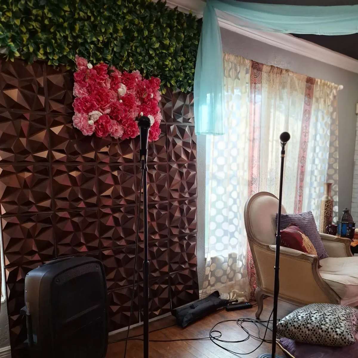 A room with two microphone stands positioned in front of a decorative wall covered in brown geometric panels and a floral arrangement, with large windows draped in sheer curtains.
