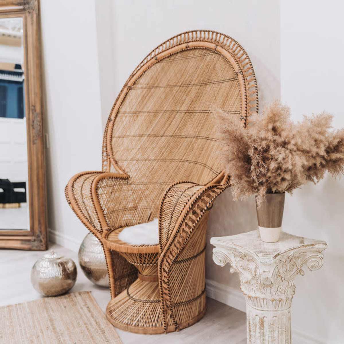 A large, woven rattan chair is positioned next to a decorative column with a vase of dried flowers, against a light-colored wall and a large mirror in the background.