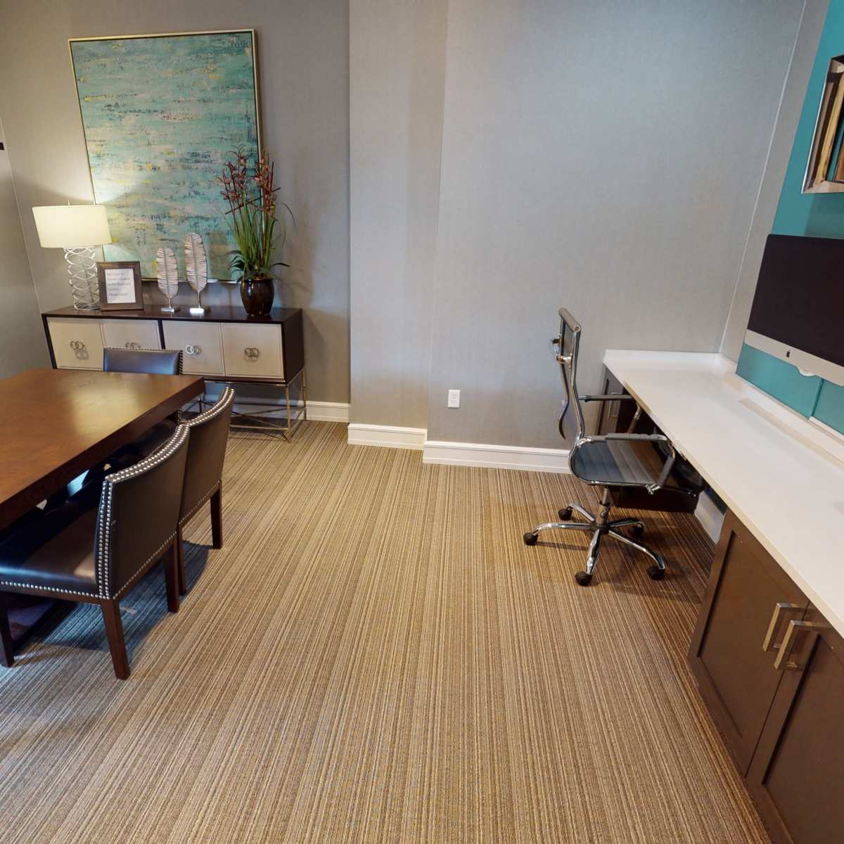 A modern office space featuring a large wooden dining table surrounded by chairs, a computer desk with an Apple monitor, and a printer nearby, all set against a neutral-toned wall.