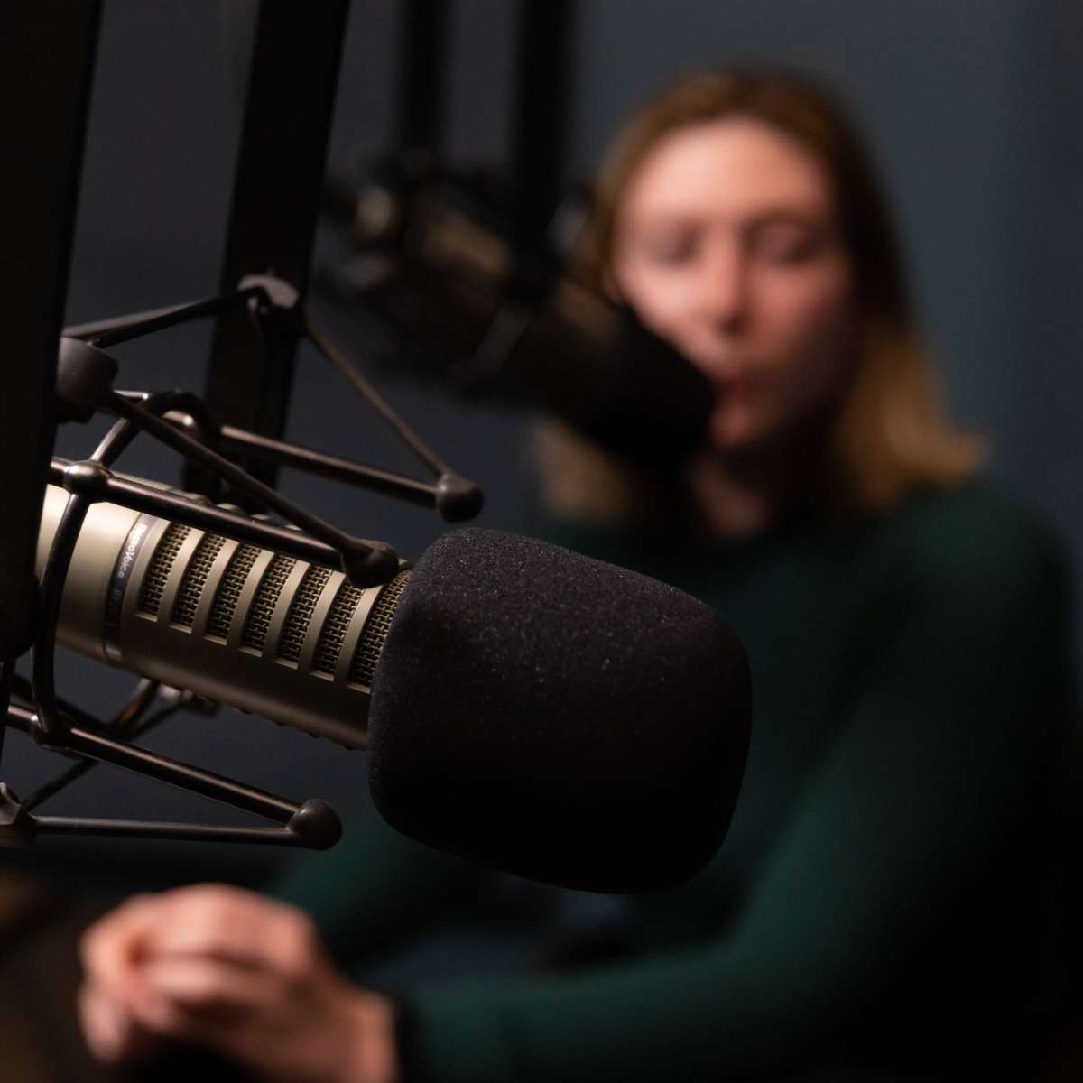 A close-up of a microphone in a recording studio with a blurred figure sitting behind it.