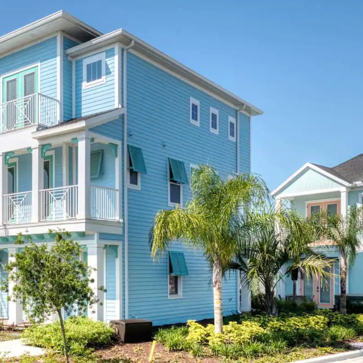 A three-story blue house with a balcony and palm trees, located among similar residential buildings in a landscaped area.