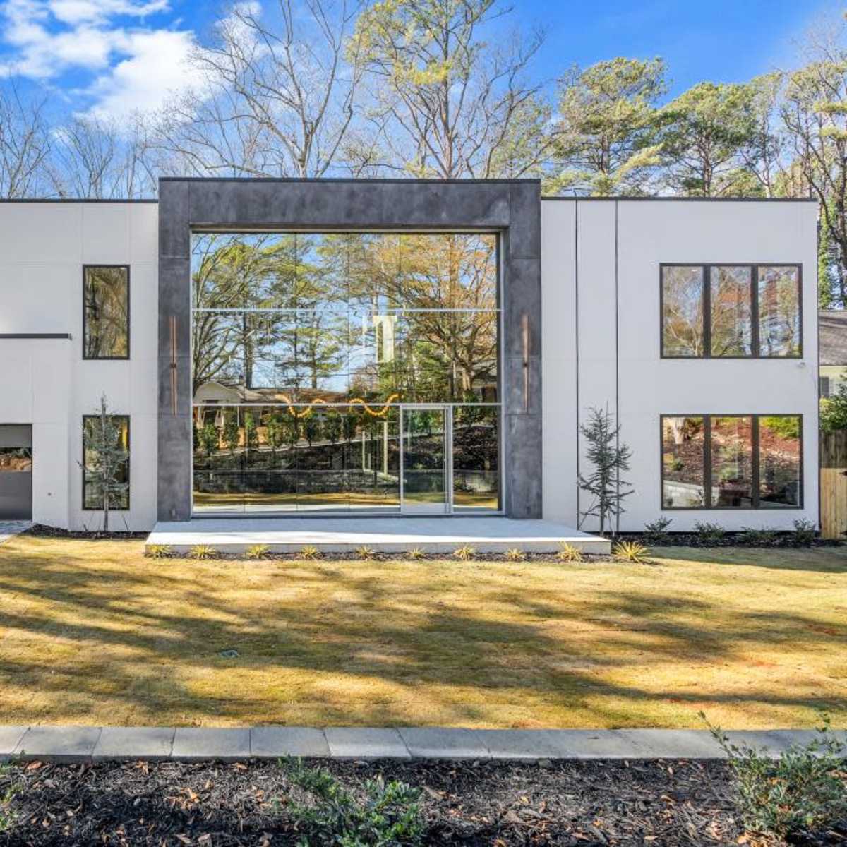 A modern two-story house with large glass windows and a landscaped front yard is set against a backdrop of trees.