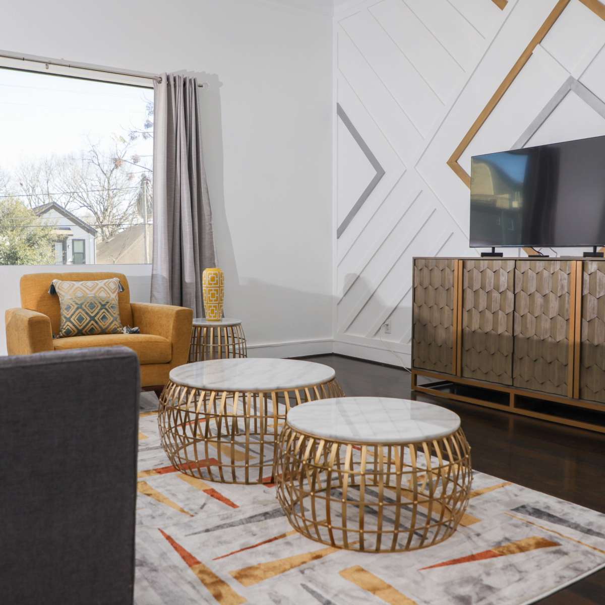 A modern living room with a gray sofa, an orange armchair, a television on a stylish cabinet, and decorative tables on a patterned rug.