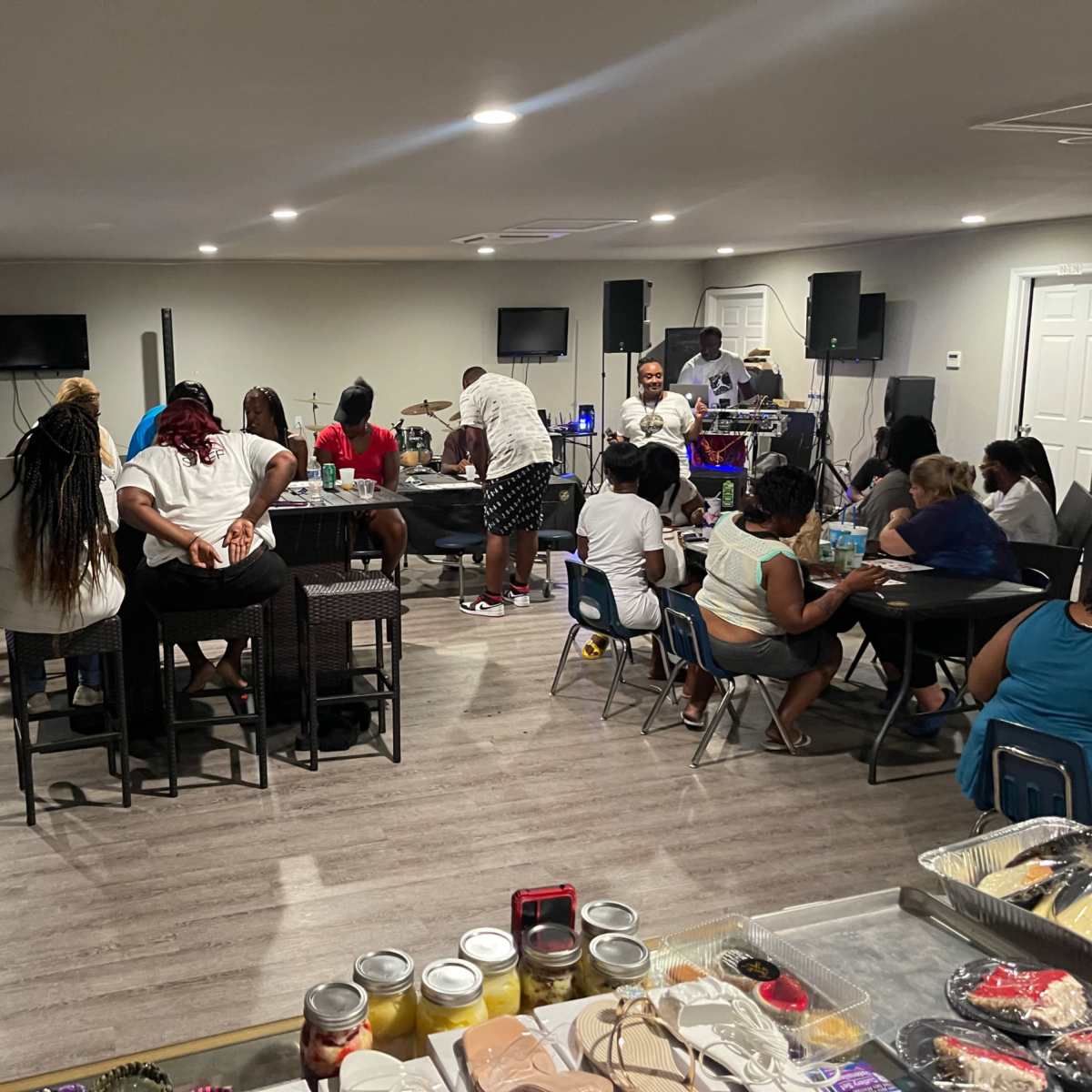 A group of people are enjoying a social gathering in a spacious room with tables and chairs, while others are performing music in the background.