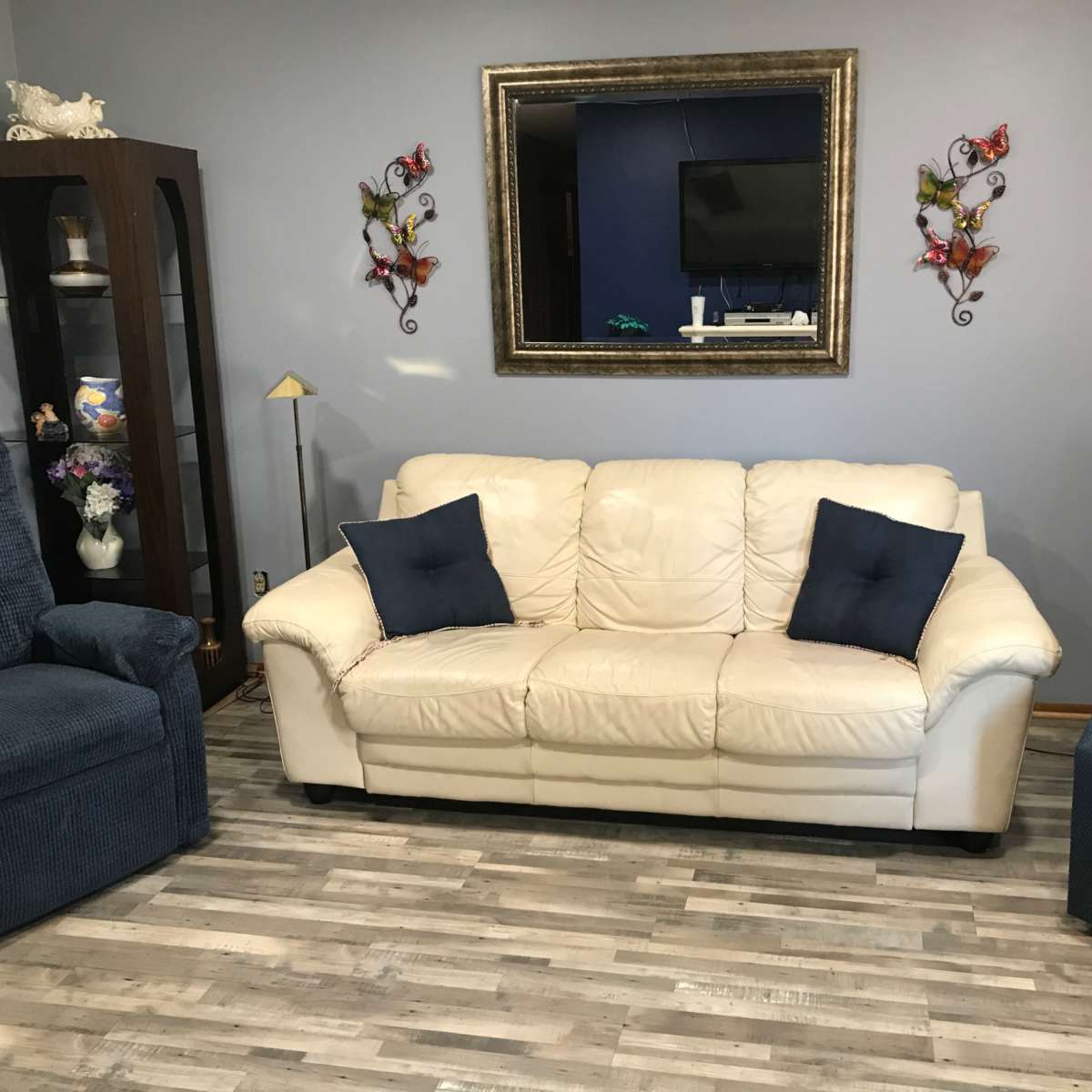 A living room with a white leather couch, two upholstered armchairs, a large mirror on the wall, and decorative items including butterflies and a lamp.