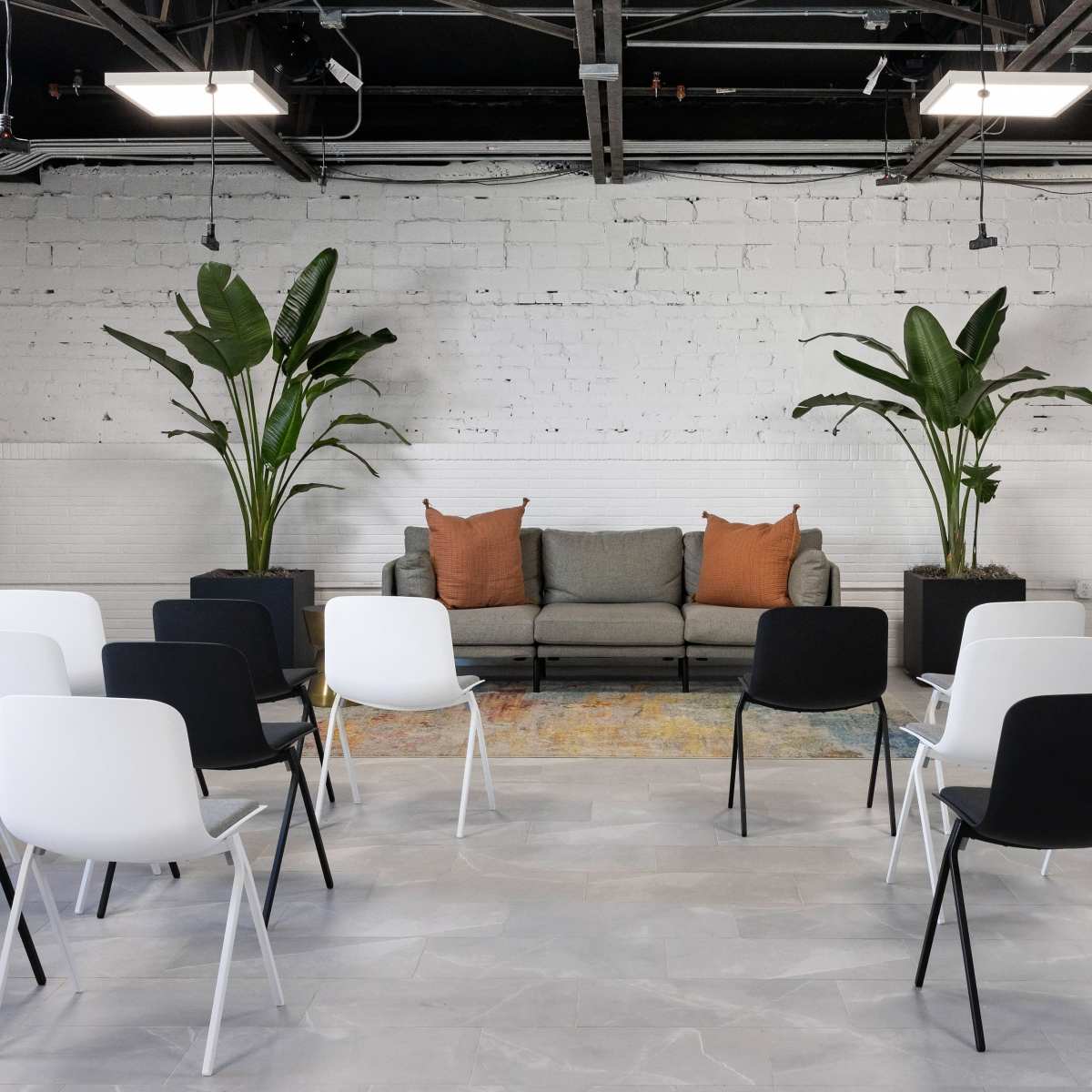 A minimalist conference room features rows of black and white chairs facing a gray sofa, flanked by potted plants against a white brick wall.