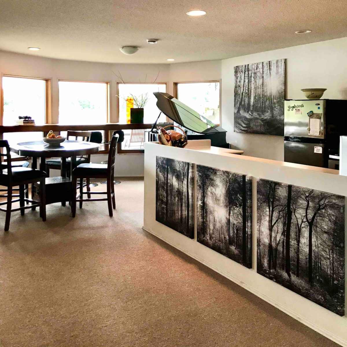 A well-lit, contemporary kitchen and dining area featuring a circular layout with a wooden dining table, chairs, and a wall decorated with black-and-white forest photographs.