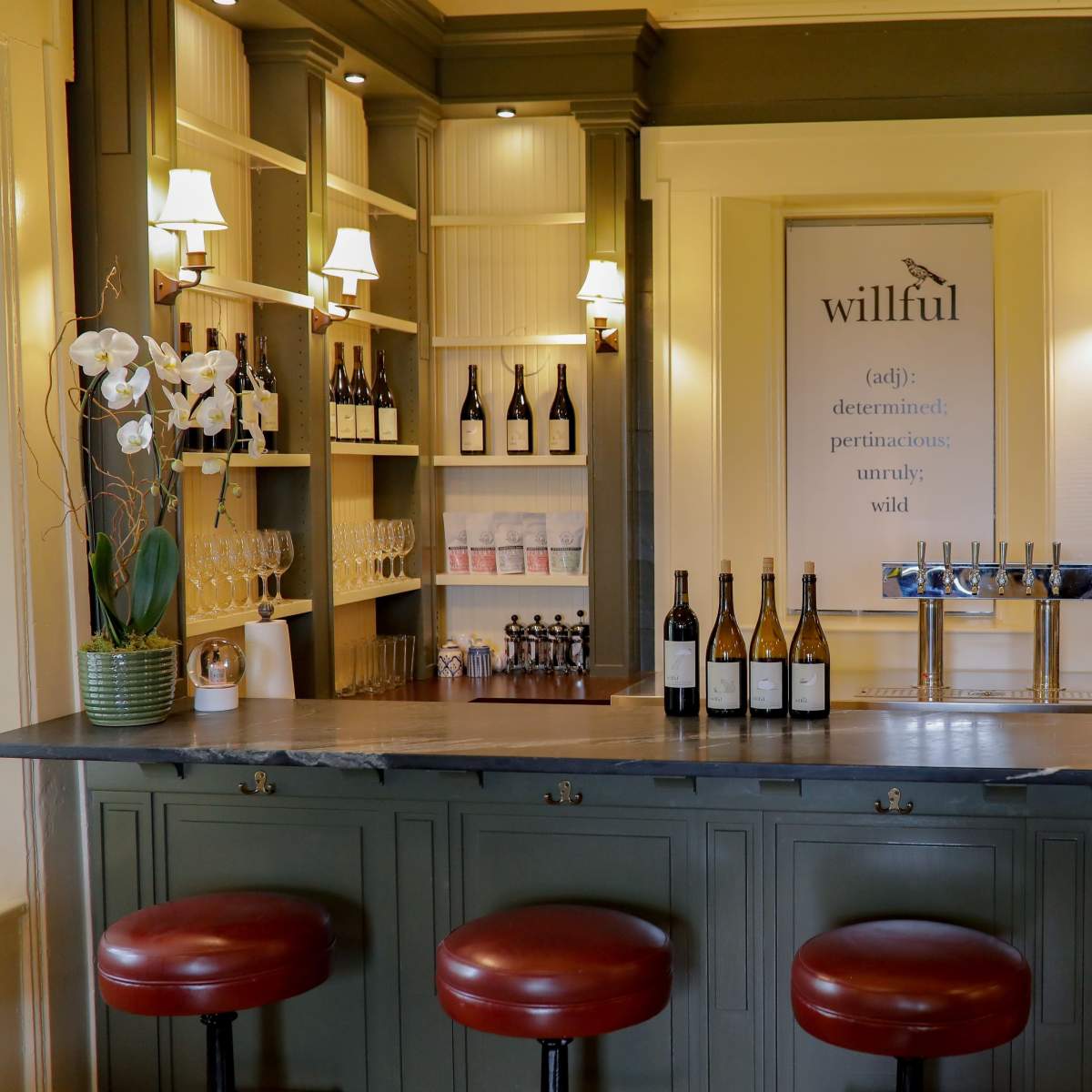 A wooden bar with three red stools displays several wine bottles and a brewing station, surrounded by shelves of decorative teapots and cups.
