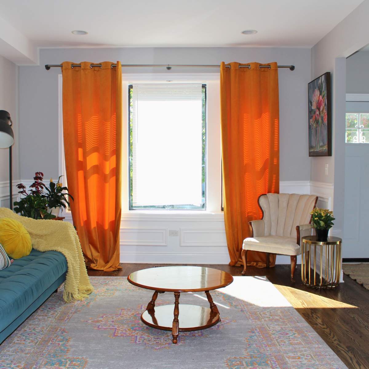 A bright living room with a blue sofa, an orange curtain over a window, and a small circular wooden coffee table.