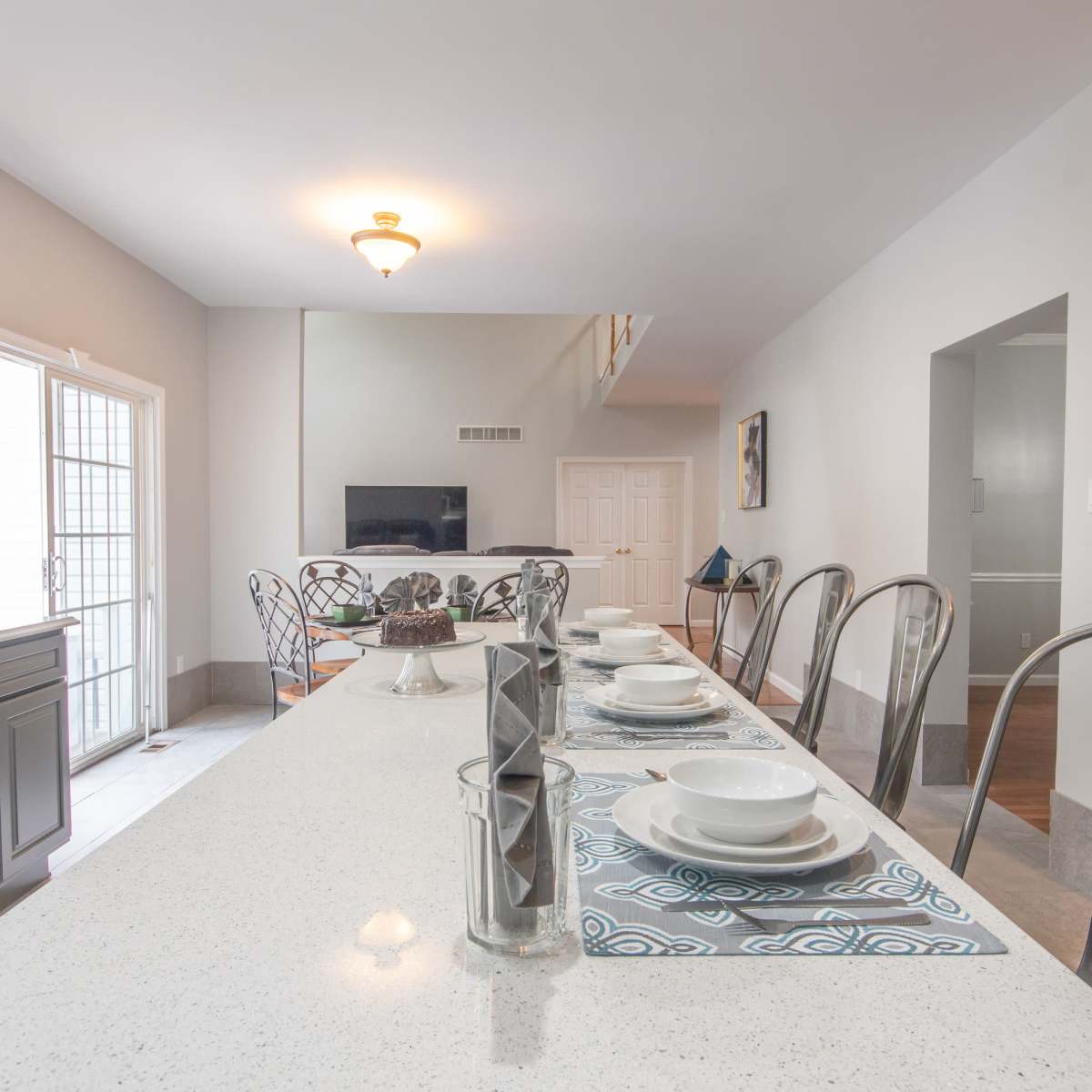 A spacious kitchen features a long countertop with set dining ware and a view of an adjoining living area with a television.