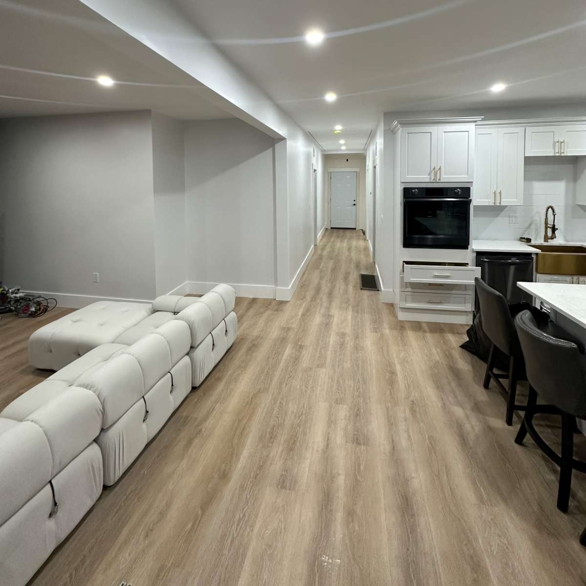 A modern, open-concept living space featuring a white sectional sofa, light-colored wood flooring, and a kitchen area with white cabinetry and a dark countertop.