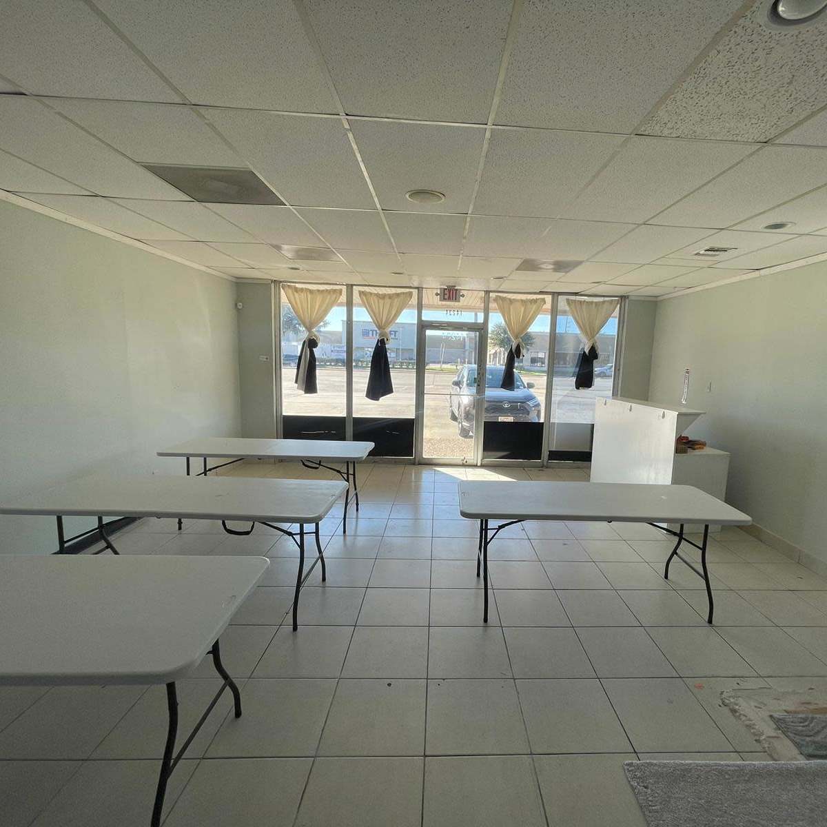 An empty room with several folding tables arranged in a grid and large windows covered with curtains.