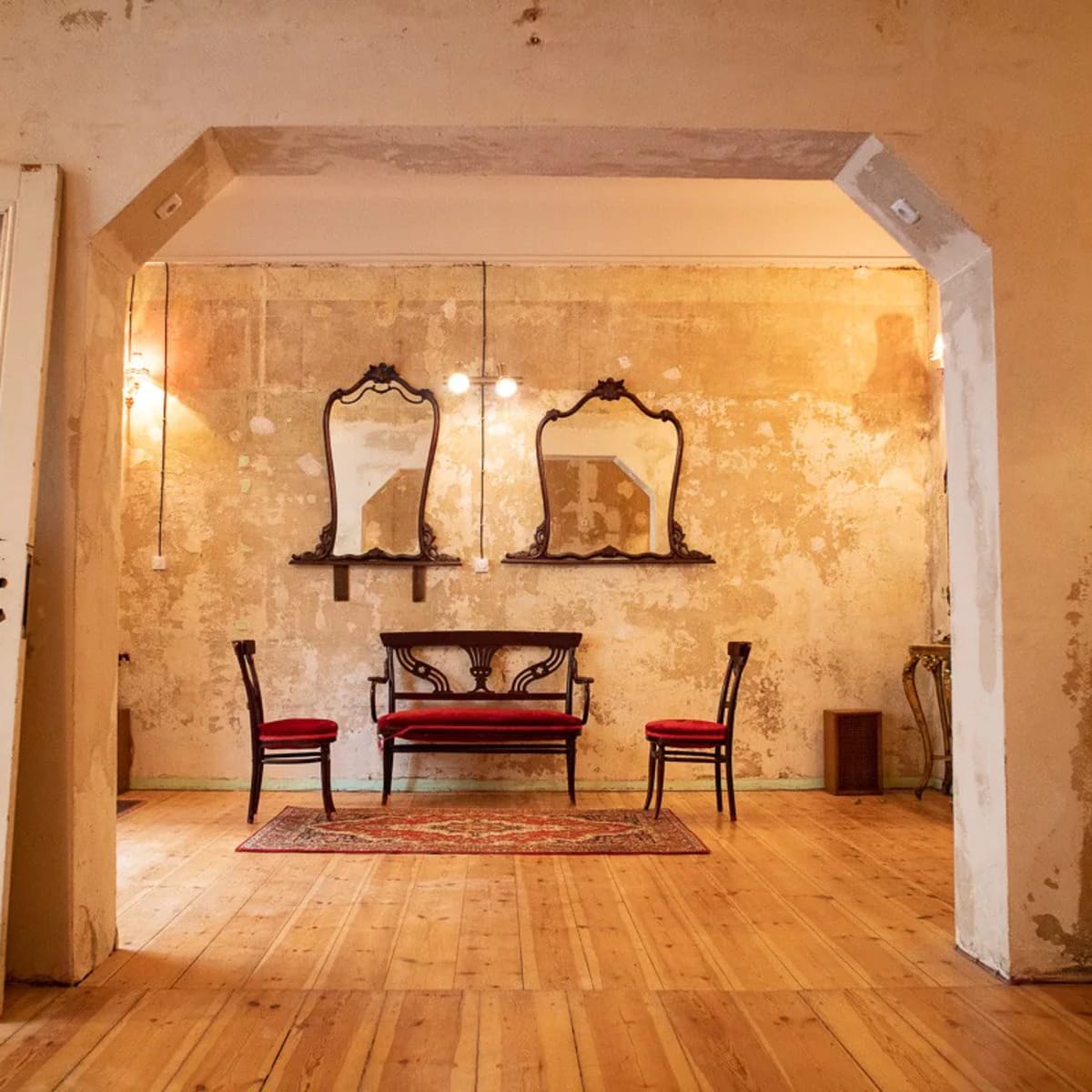A room with a wooden floor, two red chairs, a central ornate bench, and three framed mirrors on the wall, all set against a peeling plaster backdrop.