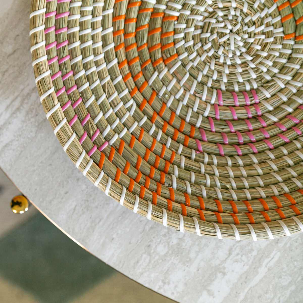 A circular woven basket with colorful accents placed on a textured surface, partially overlapping a patterned rug.