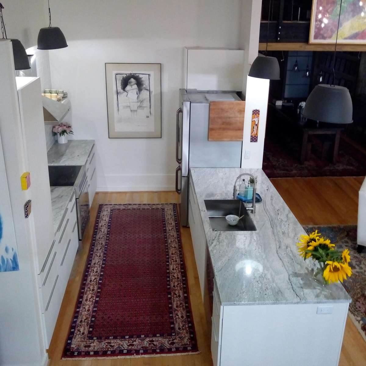 A modern kitchen with granite countertops, a stainless steel refrigerator, and a colorful rug, featuring a vase of sunflowers on the counter.