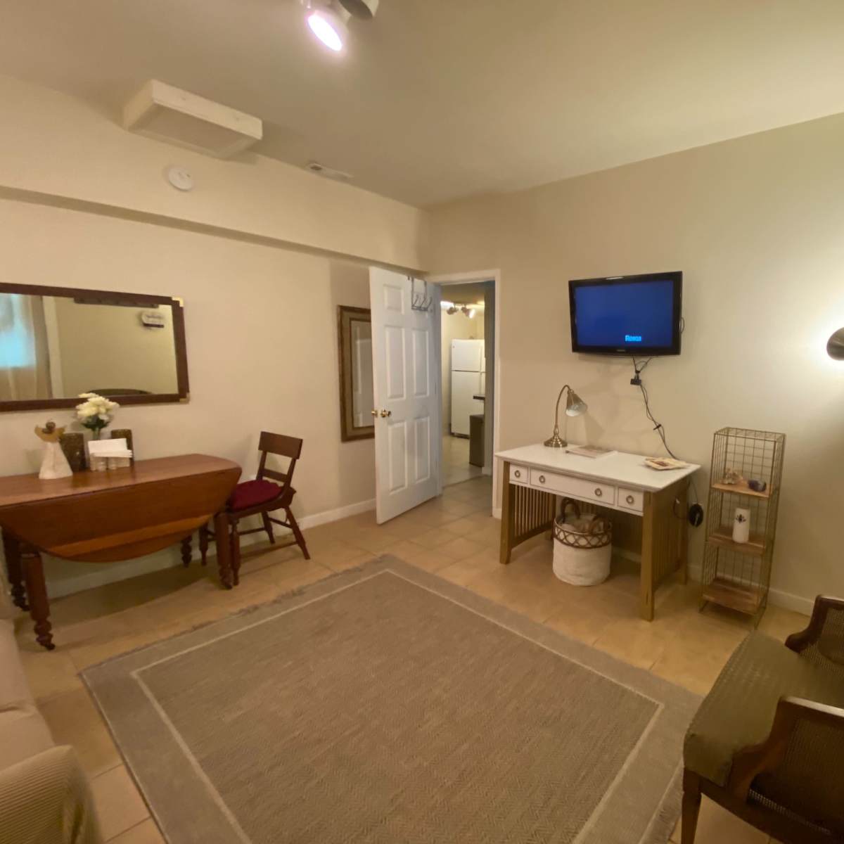 A small room furnished with a table, a chair, a mirror, a television mounted on the wall, and a rug on the tiled floor.