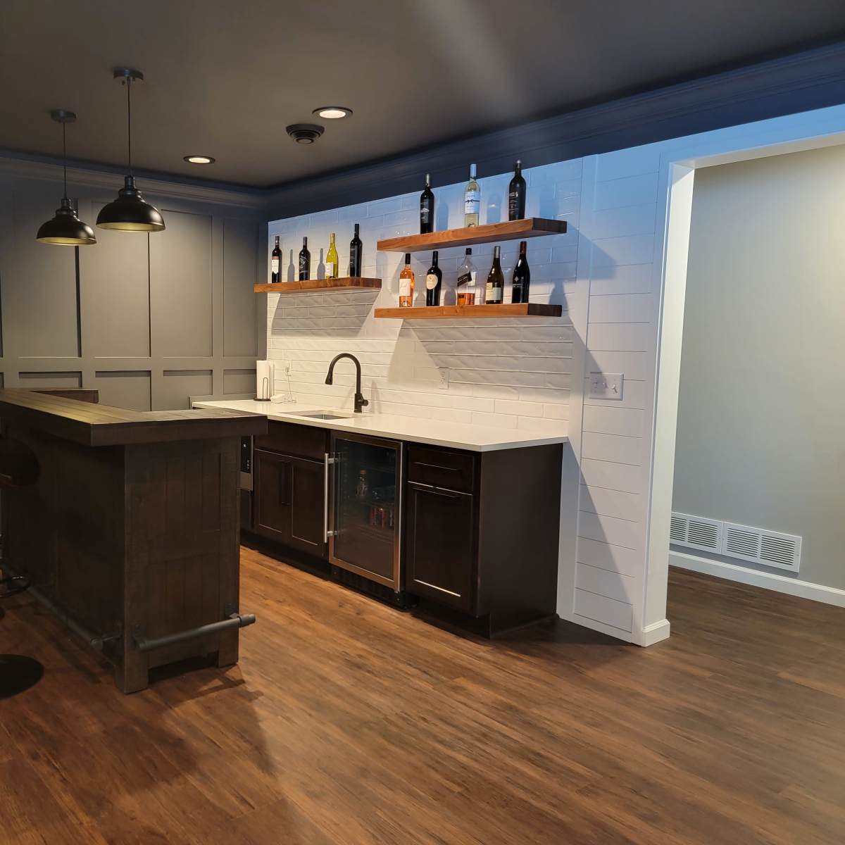 A modern kitchen bar area with a dark wood counter, two high stools, and shelves displaying various bottles against a white tiled wall.