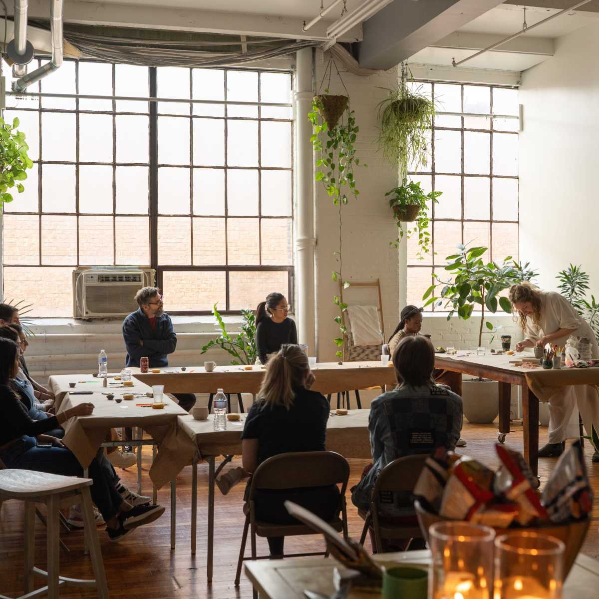 A diverse group of people gathers around a long table in a brightly lit, plant-filled room for a collaborative activity.