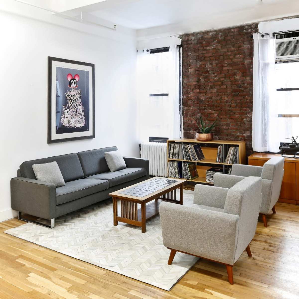 A modern living room featuring a gray sectional sofa, two gray armchairs, a wooden coffee table, a bookshelf with vinyl records, and a large framed artwork on the wall against a brick backdrop.