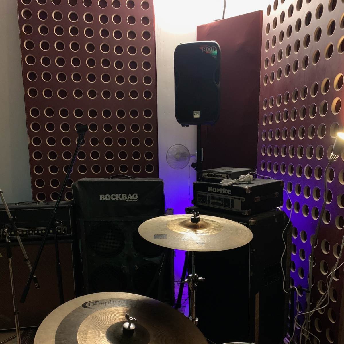 A music rehearsal space with a drum set, amplifiers, a microphone stand, and soundproofing panels on the walls.