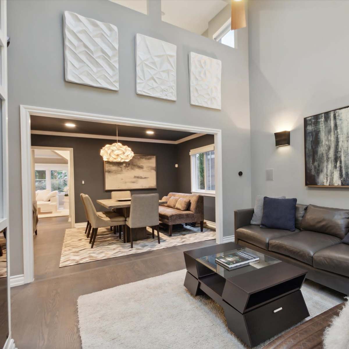A modern living room features a dark leather sofa facing a sleek coffee table, with large artwork on the walls and a dining area visible through an archway.