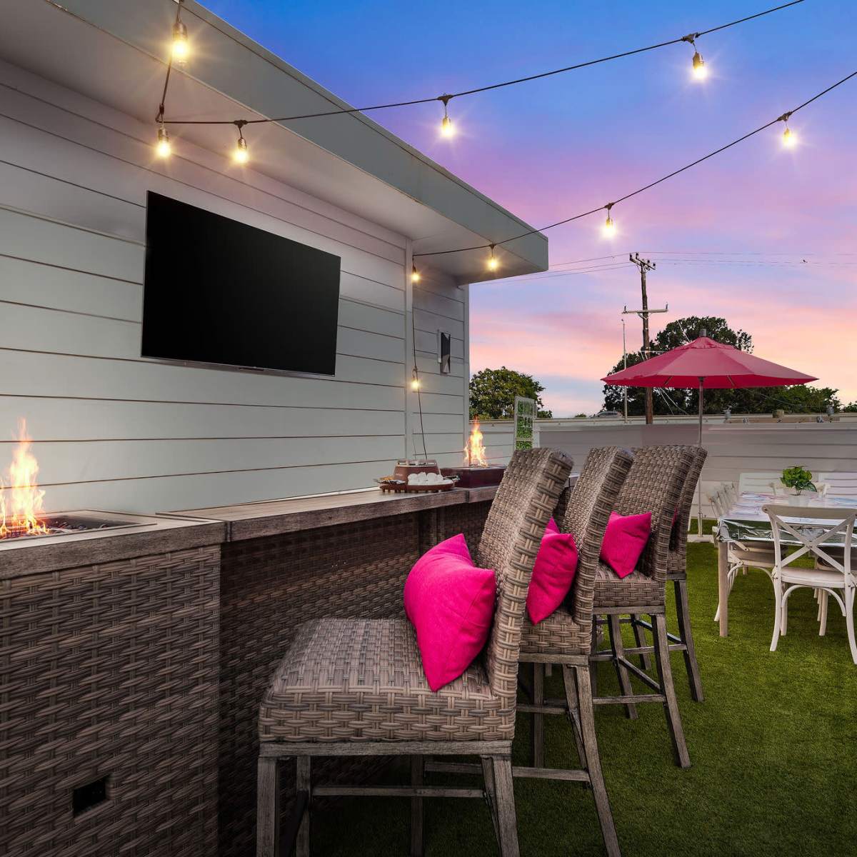A rooftop patio with a fire pit, bar seating, and a dining area, accented by string lights and a pink sunset in the background.