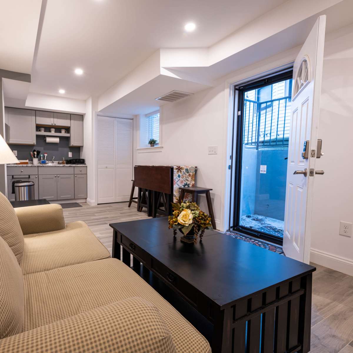 A cozy living space with a sofa, a coffee table, and a door that leads to an outdoor area, featuring a small kitchenette in the background.