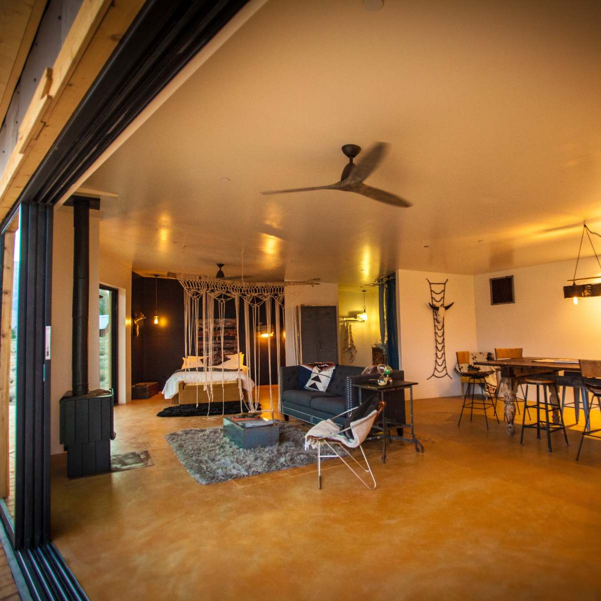 An open-concept living space featuring a dining area, a seating area, and a bedroom with a canopy bed, illuminated by pendant lighting and large windows.