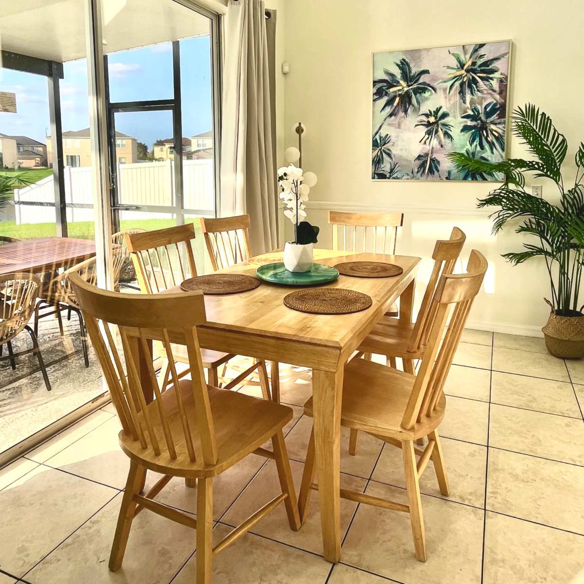 A wooden dining table surrounded by six chairs is set in a bright room, with large windows and tropical-themed art on the wall.