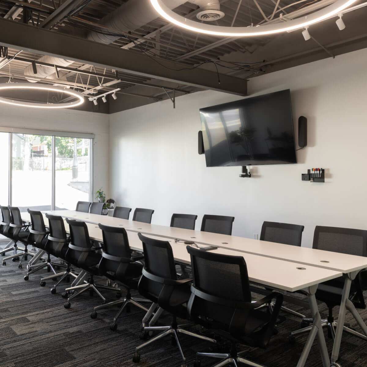 A modern conference room features a long table surrounded by black chairs, with a large flat-screen TV mounted on the wall and circular light fixtures overhead.