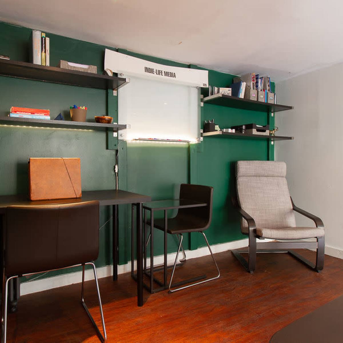 A small room with a green wall, a wooden floor, a desk with a chair, and shelves holding various items.