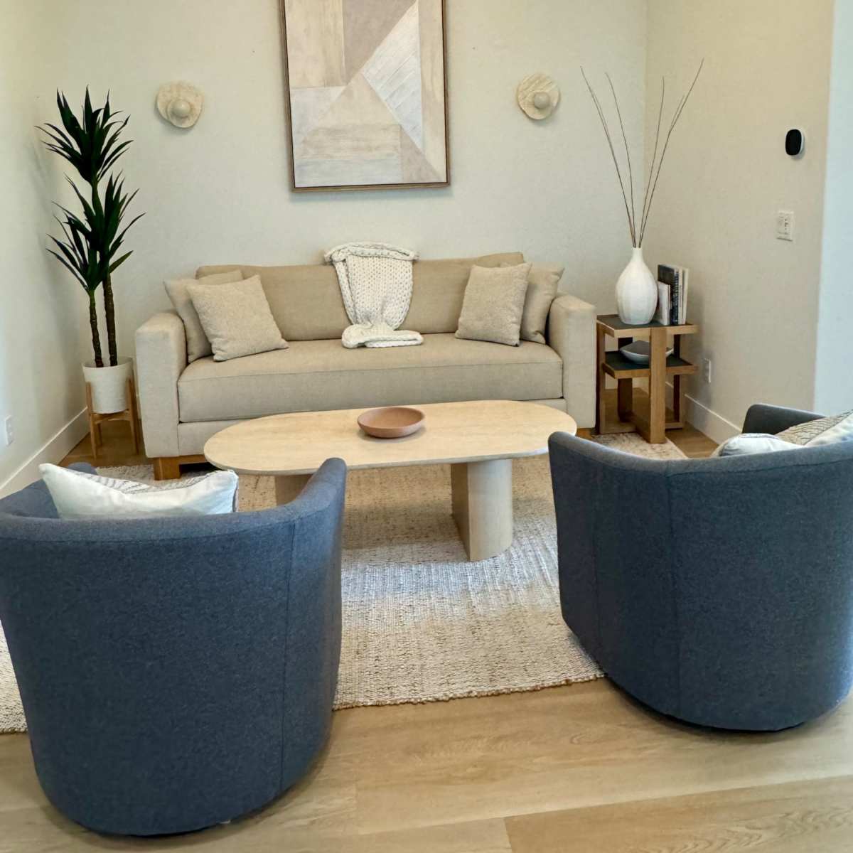 A modern living room featuring a light-colored sofa, two gray chairs, a round wooden coffee table, and decorative plants against a neutral wall.