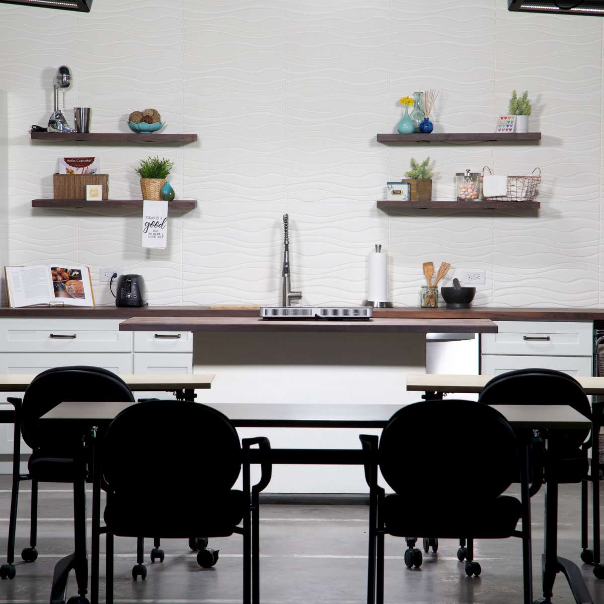 A modern kitchen setup with a cooking area, open shelving, and several appliances, positioned in front of a row of black chairs and tables.