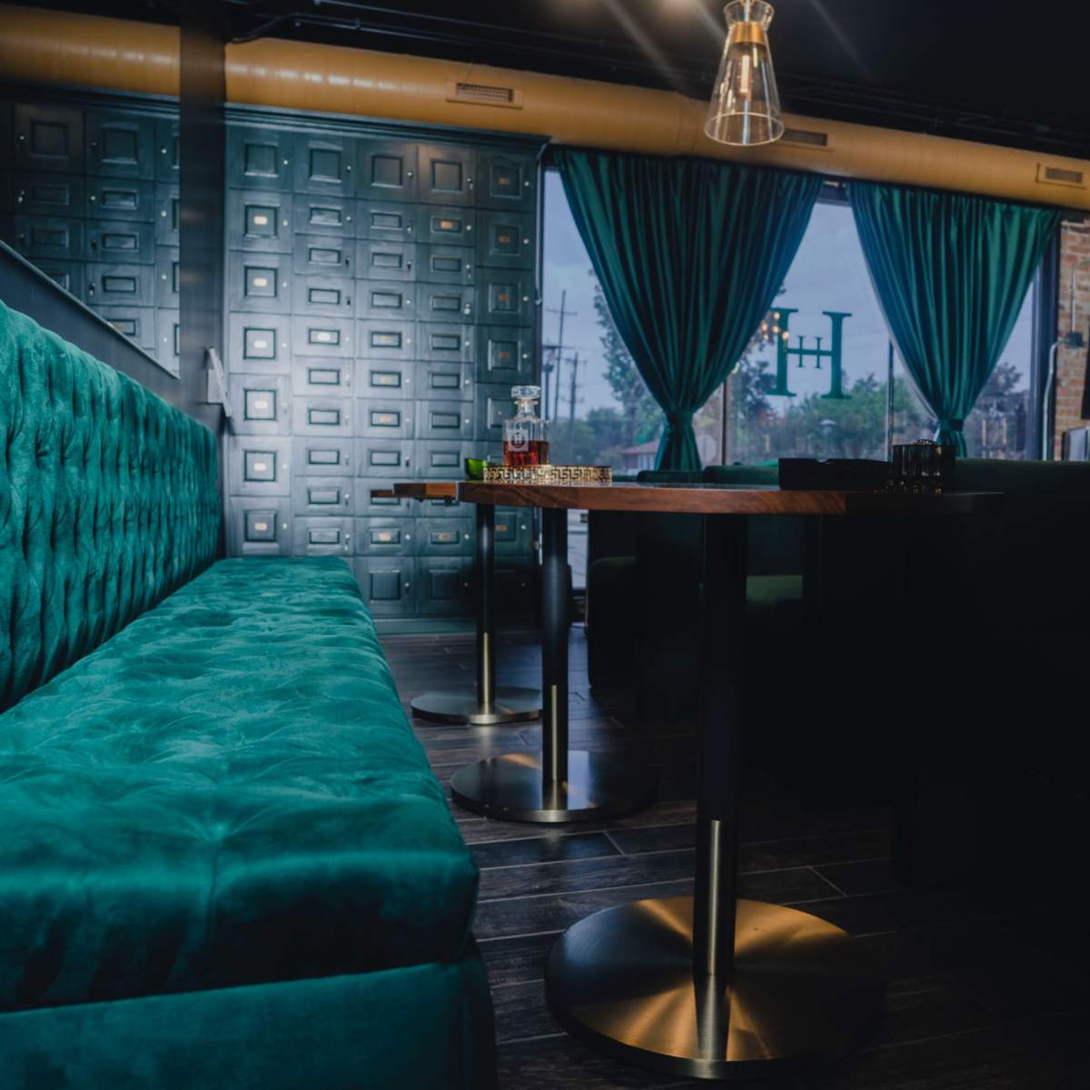 A stylish lounge area with a teal upholstered bench, a small table on a metal base, and large windows draped with teal curtains.
