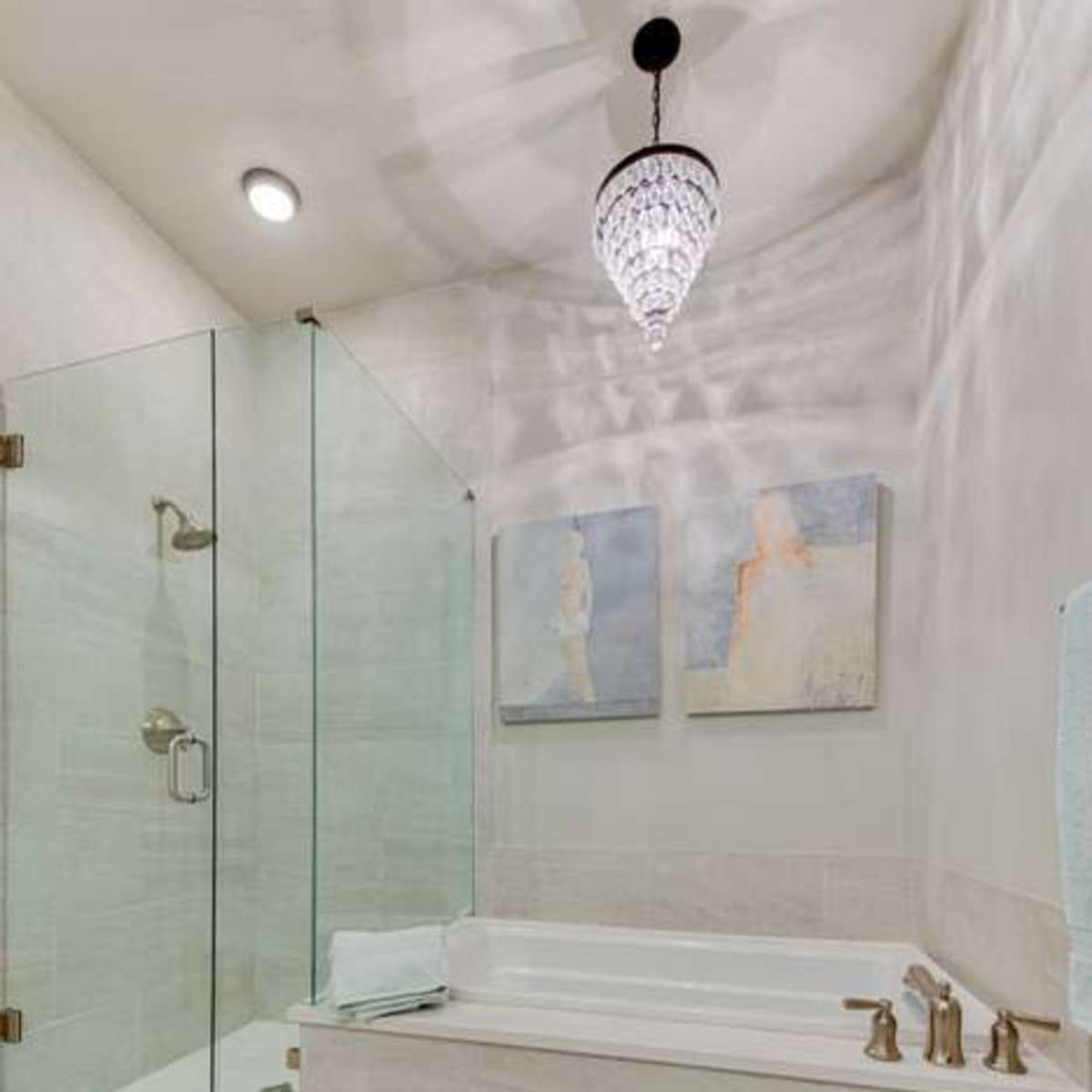 A modern bathroom featuring a glass shower enclosure, a freestanding bathtub, and wall art above the tub, illuminated by a decorative chandelier.