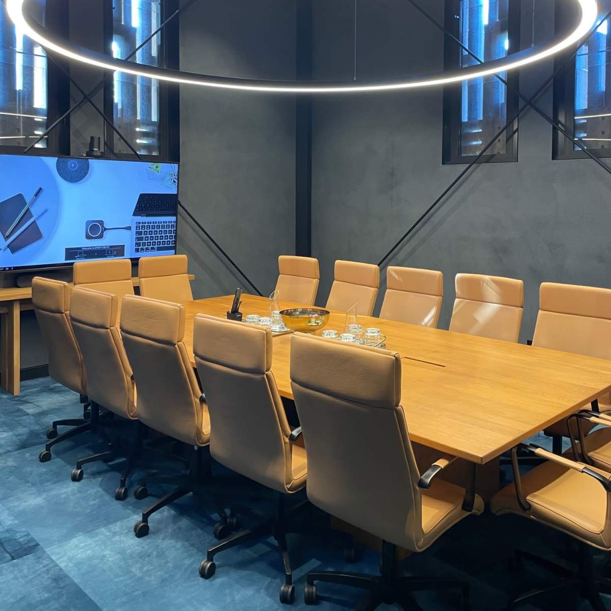 A large conference room features a rectangular wooden table surrounded by beige chairs, with a circular light fixture hanging above and a screen displaying technology-related content.