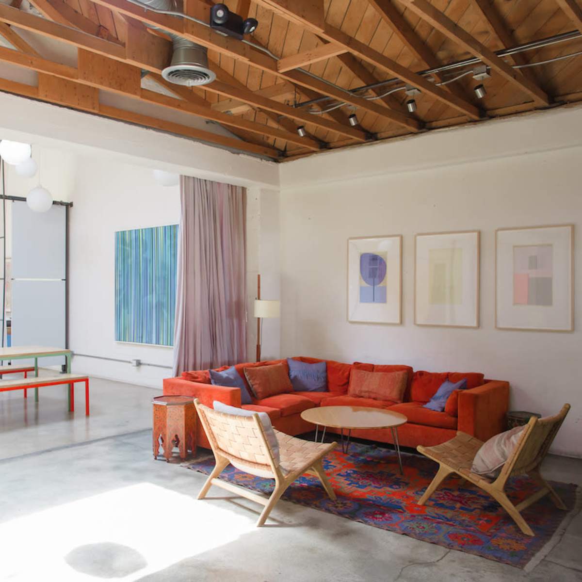 A bright, open living space featuring an orange sectional sofa, a round coffee table, and wooden chairs, with large windows and minimalist decor.