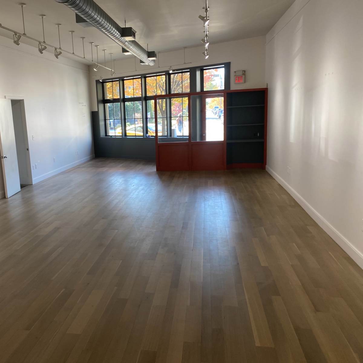 An empty room with wooden flooring, large windows, and a red bookshelf against one wall.