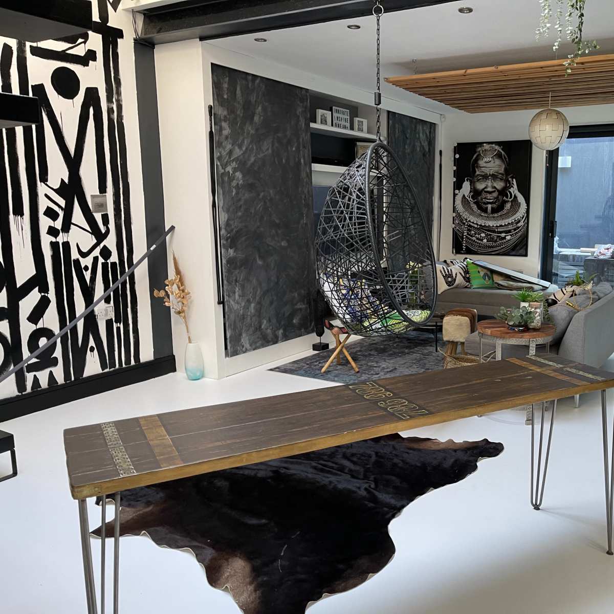 A modern living room featuring a large abstract mural, a hanging chair, a wooden table, and various furnishings including a rug and plants.