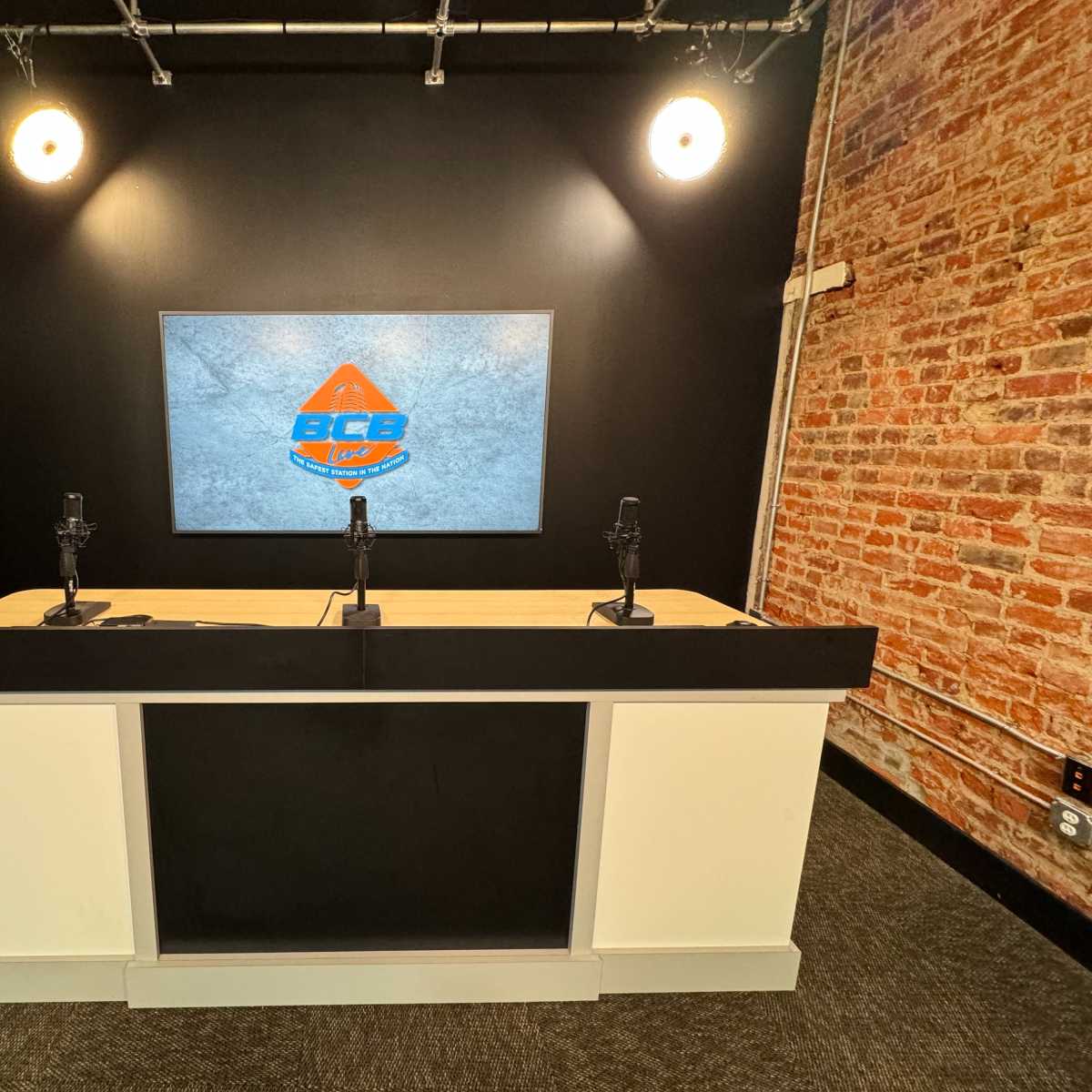 A modern broadcasting studio with a black and white counter, two microphones, and a large screen displaying a logo against a brick wall.