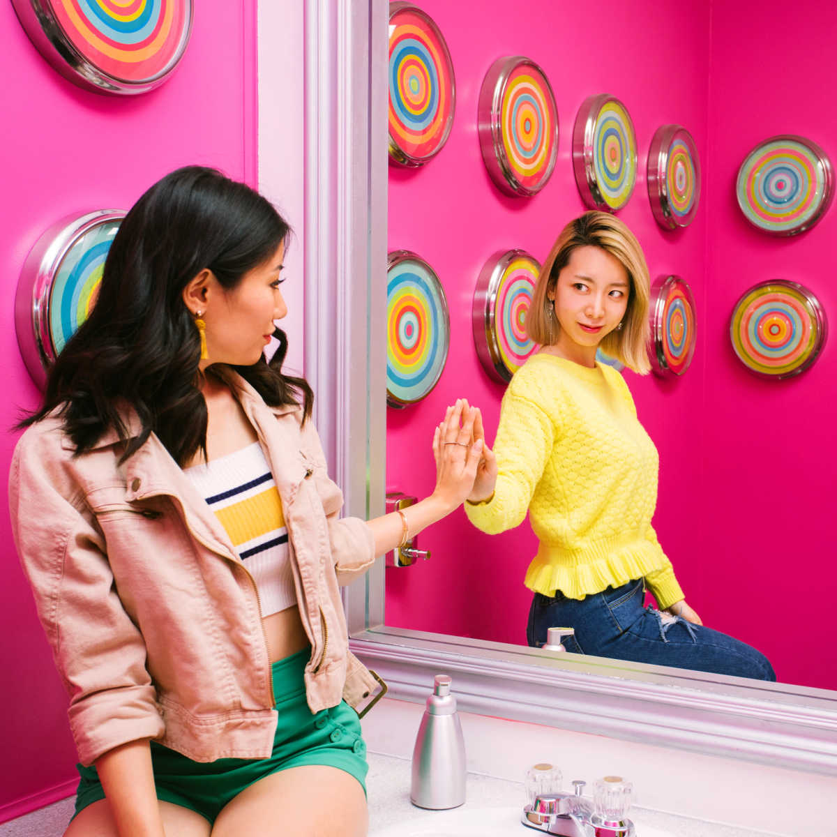 Two women are interacting in a brightly colored bathroom with a pink wall adorned with circular wall decor and a vanity mirror.