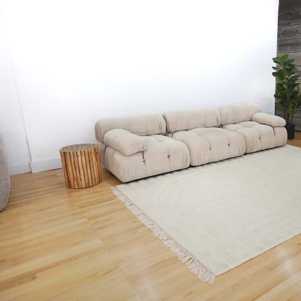 A light-colored sectional sofa and an ottoman on a large, beige rug in a spacious room with wooden flooring and a textured wall.
