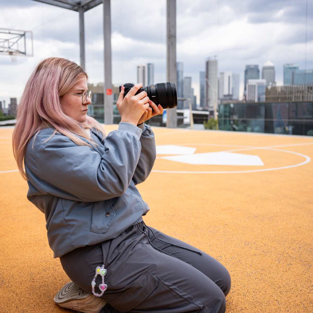 A woman with pink hair kneels on an orange basketball court while taking photos with a DSLR camera, featuring a city skyline in the background.
