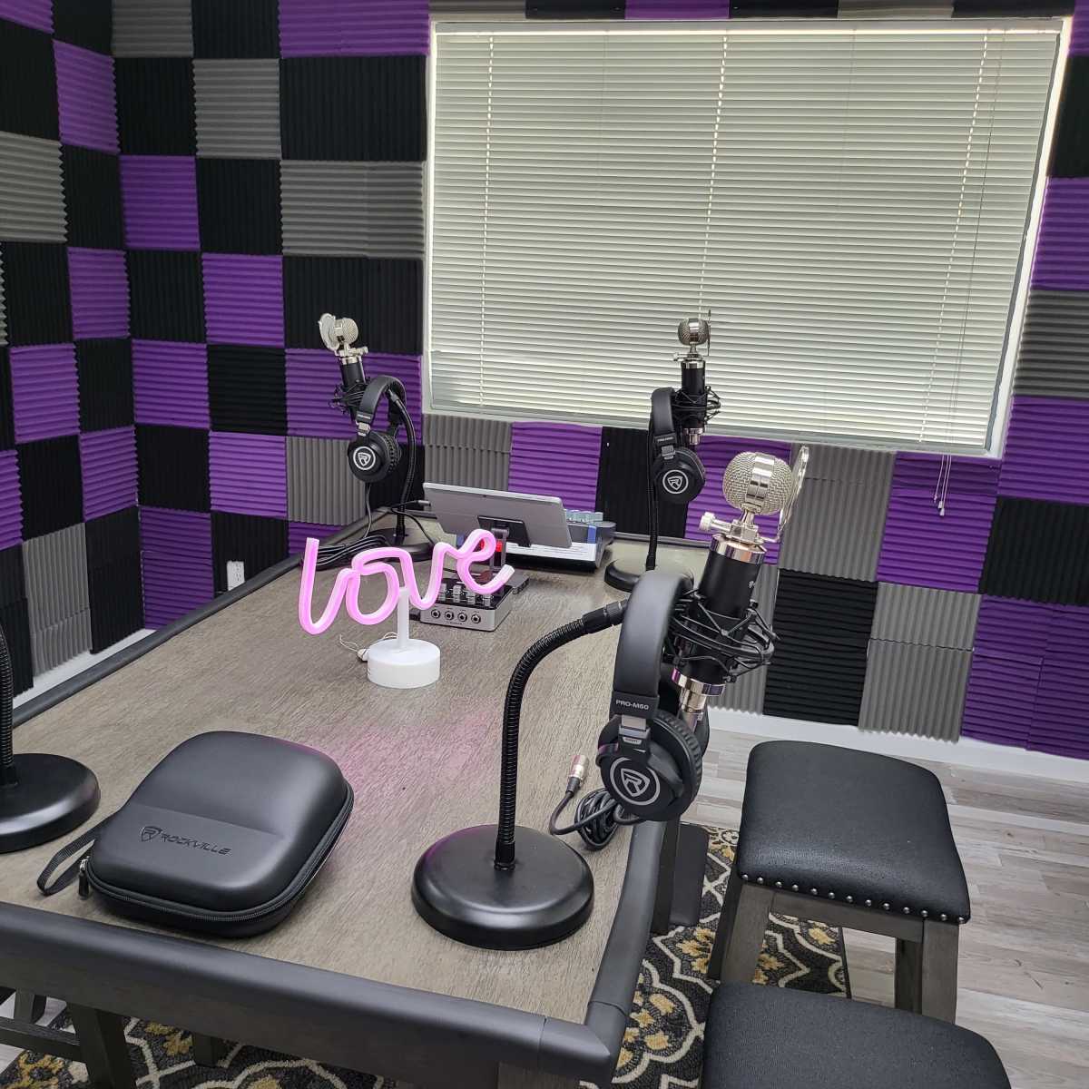 A podcast studio setup with four microphones, headphones, and a decorative "love" sign on a table, surrounded by purple and gray soundproofing panels.