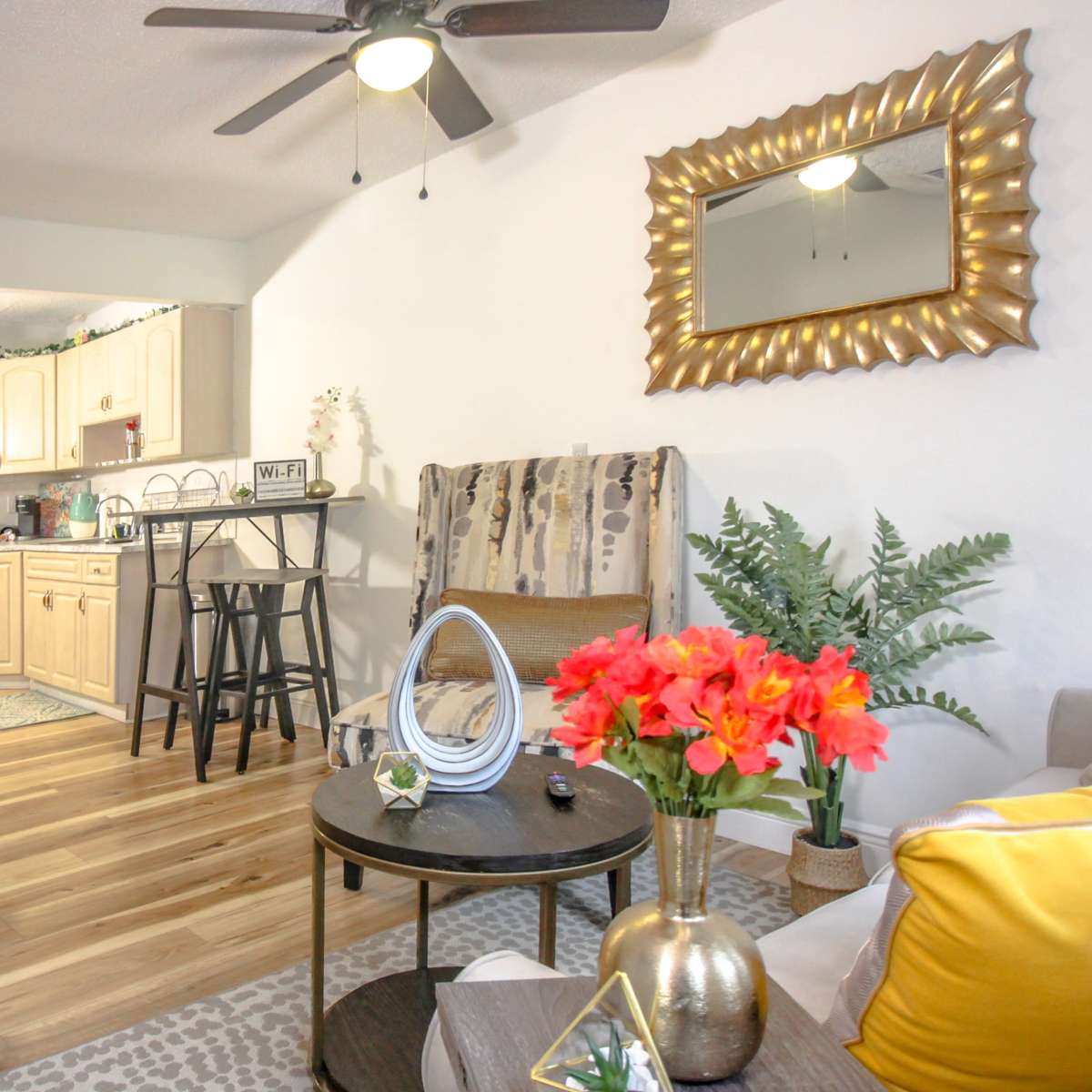 A living area with a staircase, featuring a kitchen in the background, a small dining table, and a seating area adorned with decorative plants and flowers.