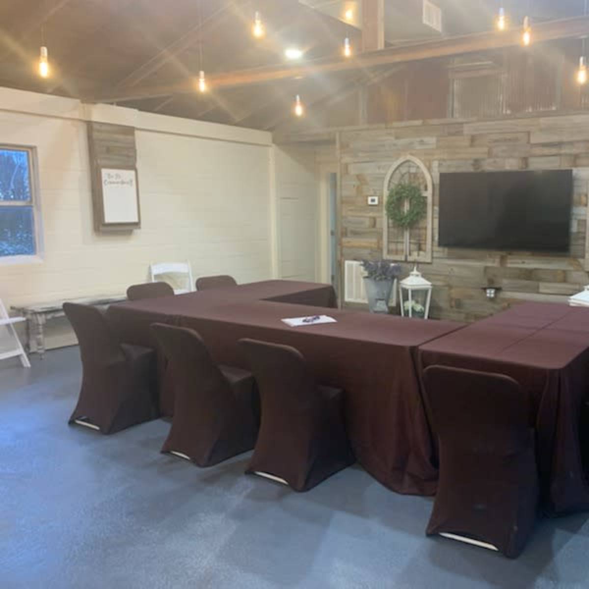 A long meeting table with dark table covers is set up in a room featuring a wooden accent wall and a large television on display.