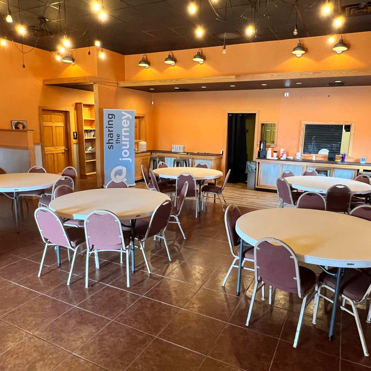 A spacious room with several round tables and chairs arranged, featuring orange walls and a service area in the background.
