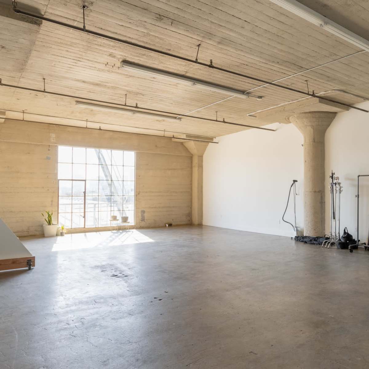 A spacious, empty room with concrete floors and large windows, featuring a few minimalist furnishings and industrial-style pillars.
