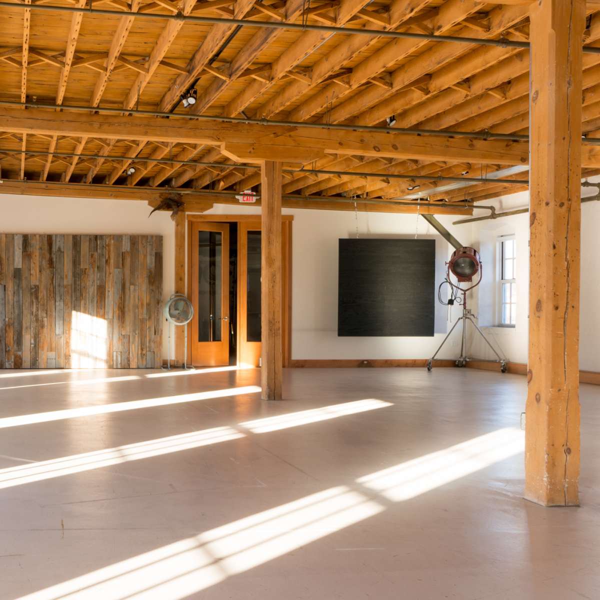 A spacious, well-lit studio with wooden beams and a polished concrete floor, featuring large windows and a wooden accent wall.